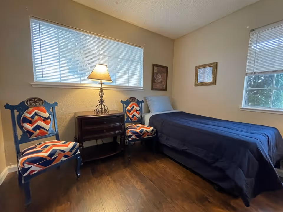 A small bedroom with a single bed covered in a dark blue blanket and a light blue pillow. Next to the bed is a dark wooden nightstand with two drawers and a decorative lamp on top. Two blue wooden chairs with colorful chevron-patterned cushions are placed beside the nightstand. The room has two windows with white blinds, beige walls, and dark wooden flooring. There are two framed pictures hanging on the walls.