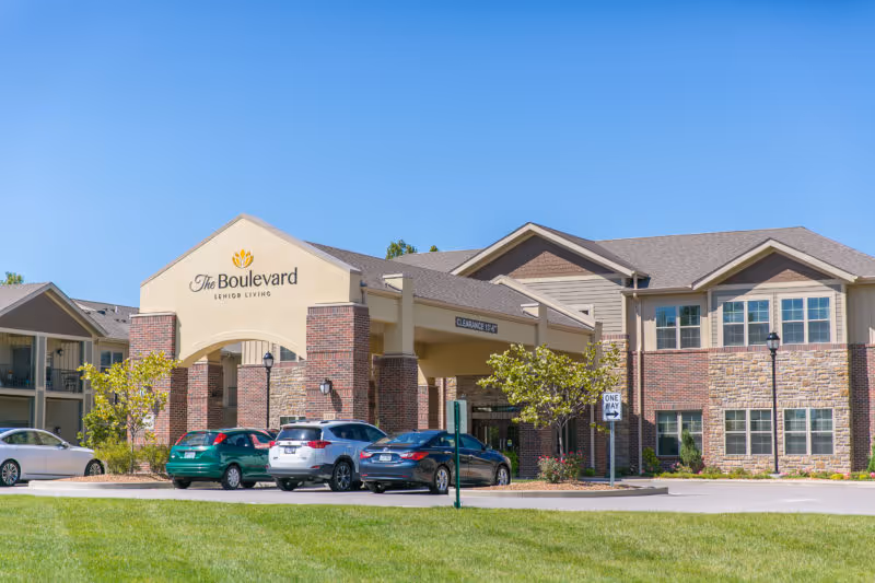 Front exterior of The Boulevard Senior Living building with a covered entrance, parked cars, and a grassy lawn under a clear blue sky.