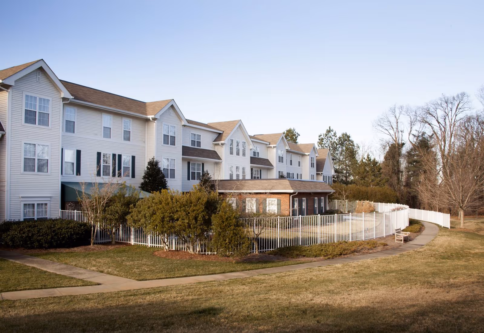 Large multi-story white senior living building with a fenced courtyard, pathway and surrounding lawn and trees.