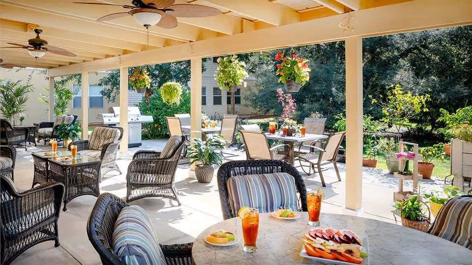 Covered outdoor patio area with wicker chairs and tables set with iced tea and plates of fruit. Hanging plants and potted plants decorate the space, with a grill and additional seating visible in the background. The area overlooks a garden with trees and greenery.