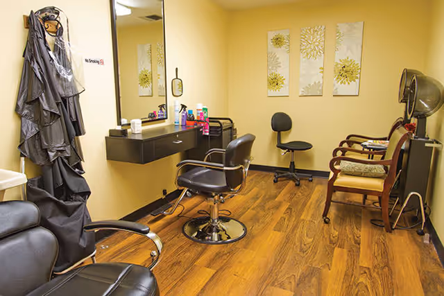 Small salon room with styling chairs, a wall mirror, hair products on a counter, and a hooded dryer on wood flooring.