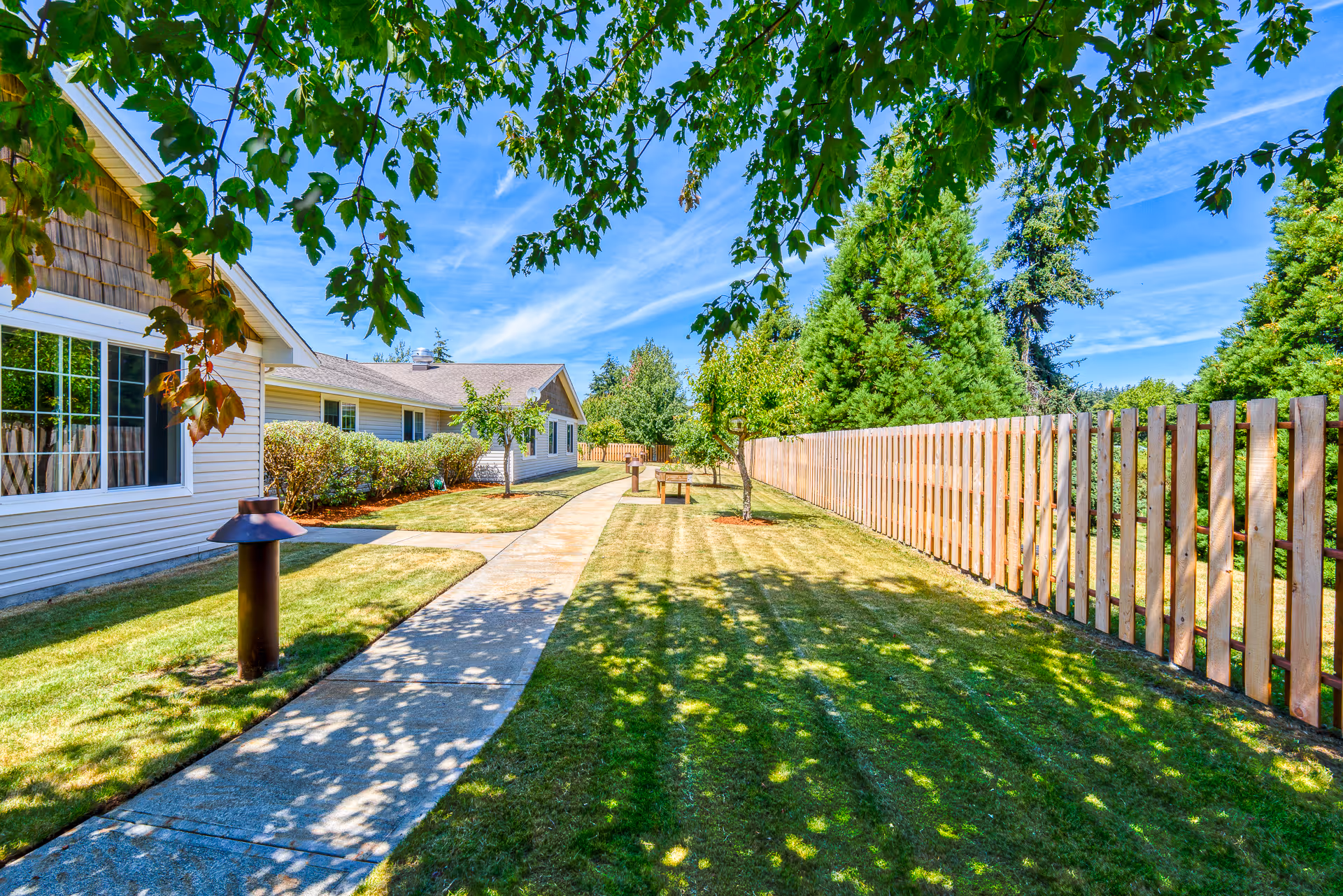 Sunlit walkway beside a single-story memory care building with manicured lawn, trees, a wooden fence, and benches.