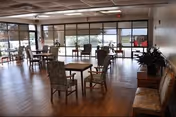 Open communal dining room with multiple tables and chairs, large floor-to-ceiling windows, wood flooring, and lounge seating along the wall.