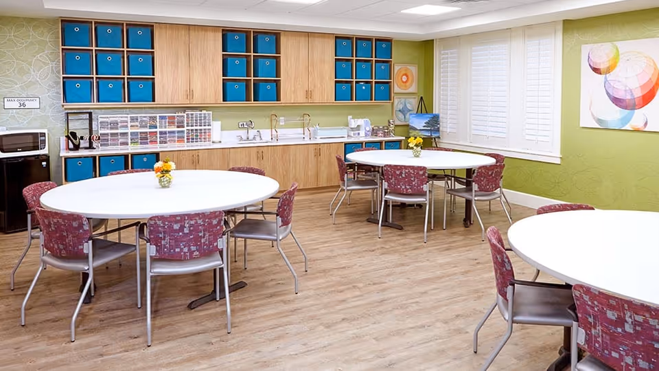 Bright multipurpose dining and activity room with round tables and chairs, cabinetry and sinks along the back wall.