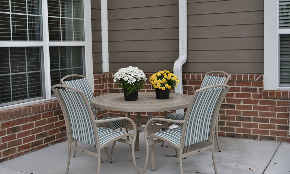 Outdoor patio area with a round metal table and four striped cushioned chairs. Two potted plants with white and yellow flowers are placed on the table. The patio is adjacent to a building with brick and siding walls and windows with white blinds.
