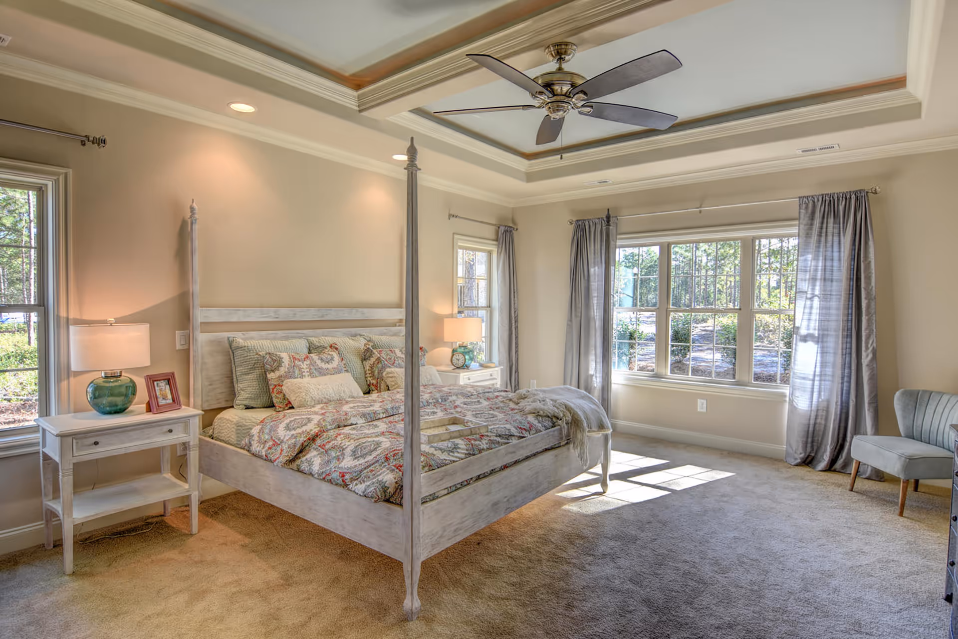 A bright and spacious bedroom with a large four-poster bed featuring patterned bedding. There are two windows with gray curtains allowing natural light to fill the room. On either side of the bed are white nightstands with lamps and decorative items. The room has beige walls, a ceiling fan, and carpeted flooring.