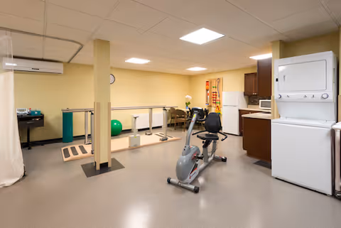 A spacious room with exercise equipment including a stationary recumbent bike and parallel bars for walking support. The room has beige walls, a white ceiling with fluorescent lights, a stacked washer and dryer, and a small kitchenette area with cabinets and a microwave. There is also a green exercise ball and a clock on the wall.