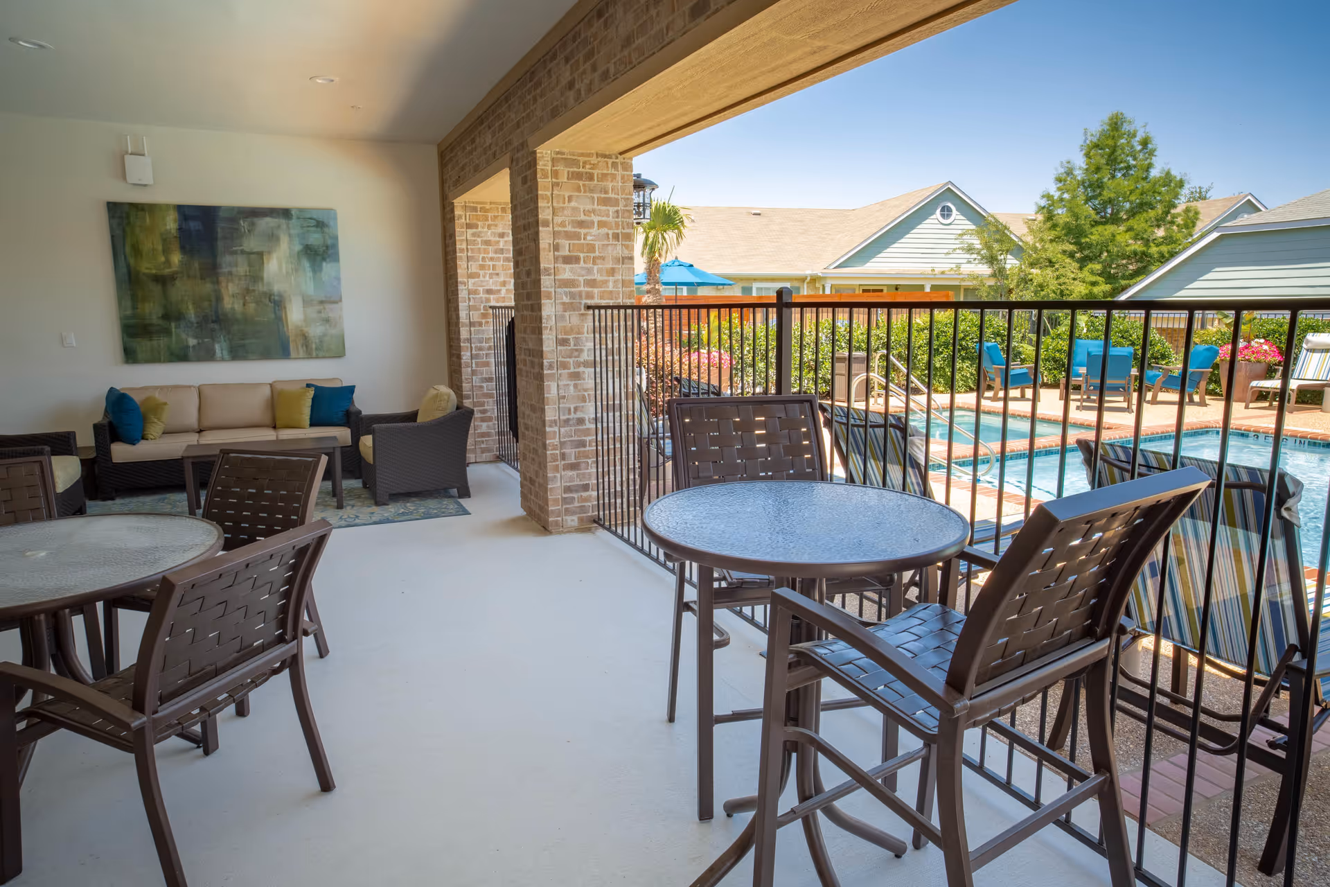 Covered patio area with outdoor seating including tables and chairs, overlooking a fenced swimming pool with lounge chairs and umbrellas. In the background, there are houses and trees under a clear blue sky.
