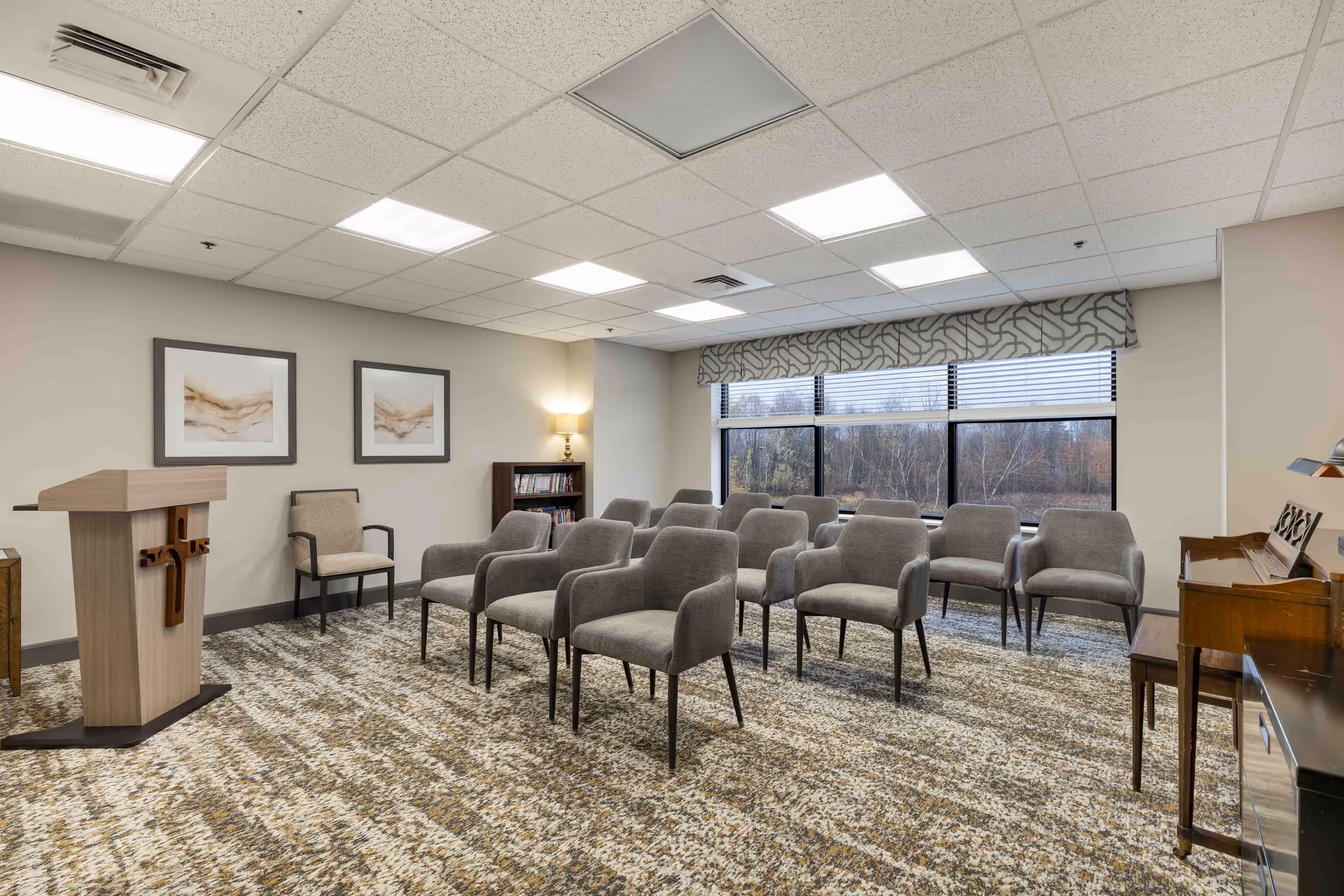 A small meeting or chapel room with rows of gray upholstered chairs arranged facing a wooden podium with a cross on it. The room has a patterned carpet, two framed abstract artworks on the wall, a bookshelf with books, a floor lamp, large windows with blinds, and a piano on the right side.