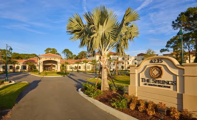 Front entrance and driveway of a Mediterranean-style senior living building with palm trees and a stone sign.