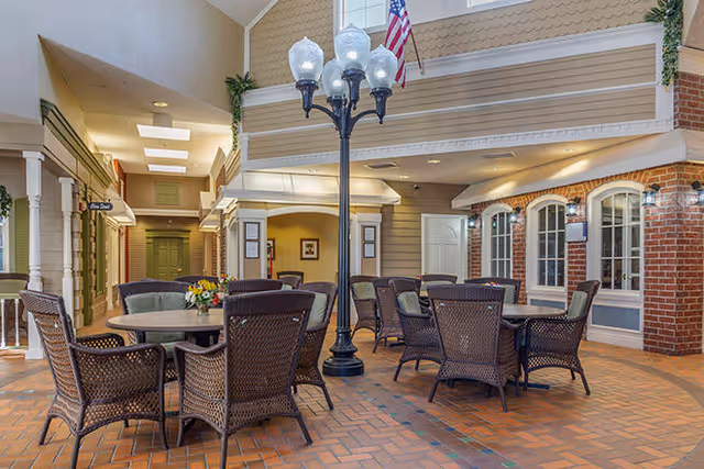 Indoor common area with multiple round tables surrounded by wicker chairs, a tall black streetlamp-style light fixture in the center, brick and siding walls with windows, and an American flag hanging from the upper level.