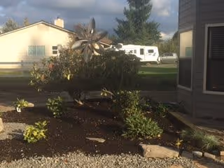 A landscaped garden area with various plants and shrubs, a decorative metal wind spinner, and a gravel pathway. In the background, there is a light-colored house, a white RV, and a large evergreen tree under a partly cloudy sky.
