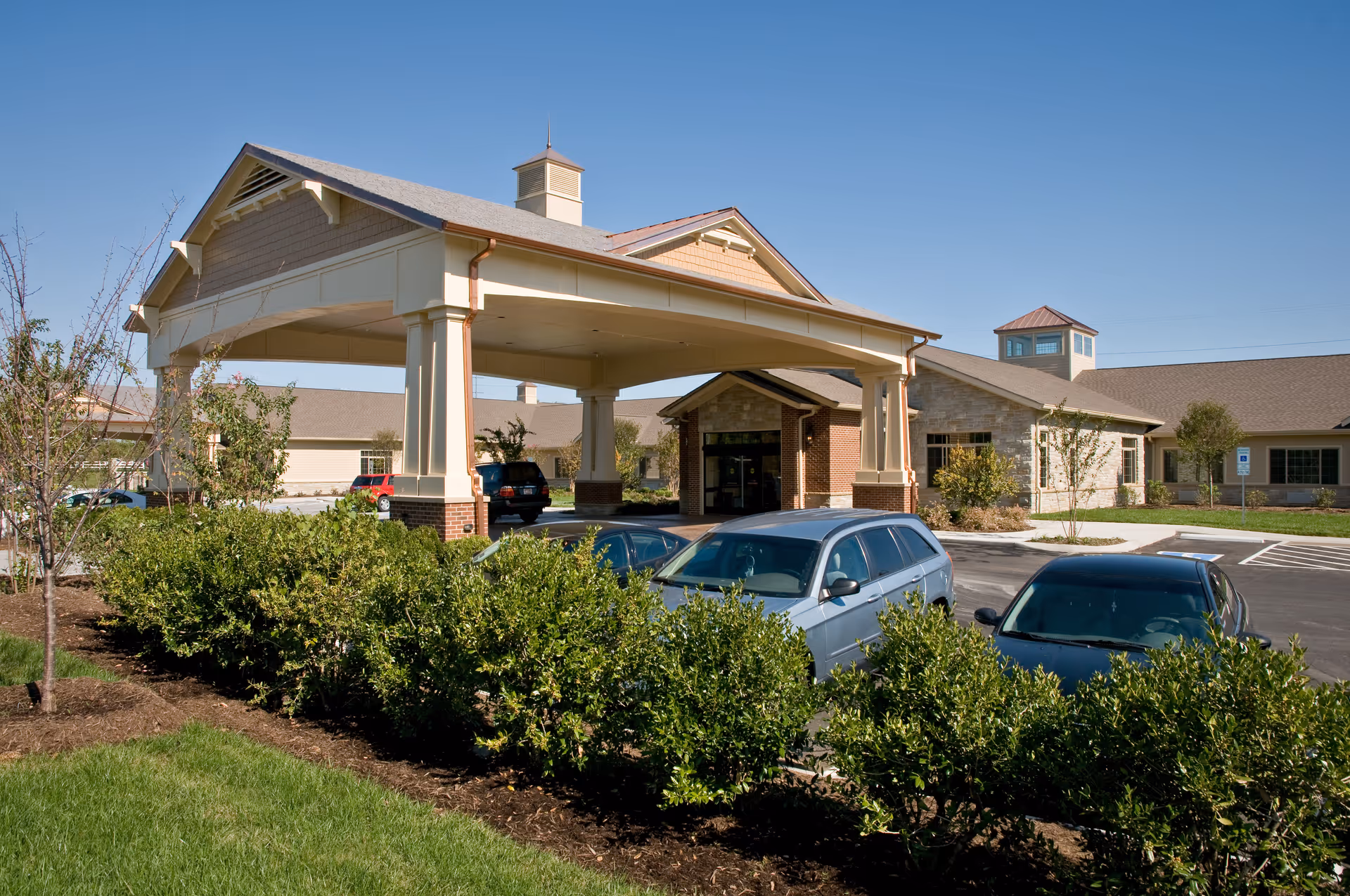 Front entrance of a senior living facility with a covered porte-cochere, parked cars, and landscaped shrubs under a clear blue sky.