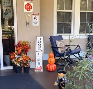 Front porch area of a building with a glass door and window. There are autumn-themed decorations including potted flowers, a vertical sign that reads 'HOME sweet HOME', three stacked pumpkins, a cushioned chair with a decorative pillow, and a small scarecrow figure. Several signs are posted on the wall near the door.