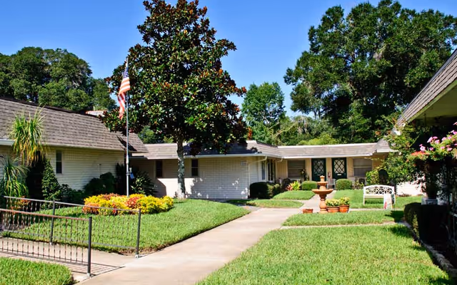 A sunny outdoor courtyard area of a senior living facility with a concrete walkway, green grass, bushes, a large tree, an American flag on a flagpole, a fountain, and a white bench. The building is single-story with white walls and a shingled roof, surrounded by trees and plants.