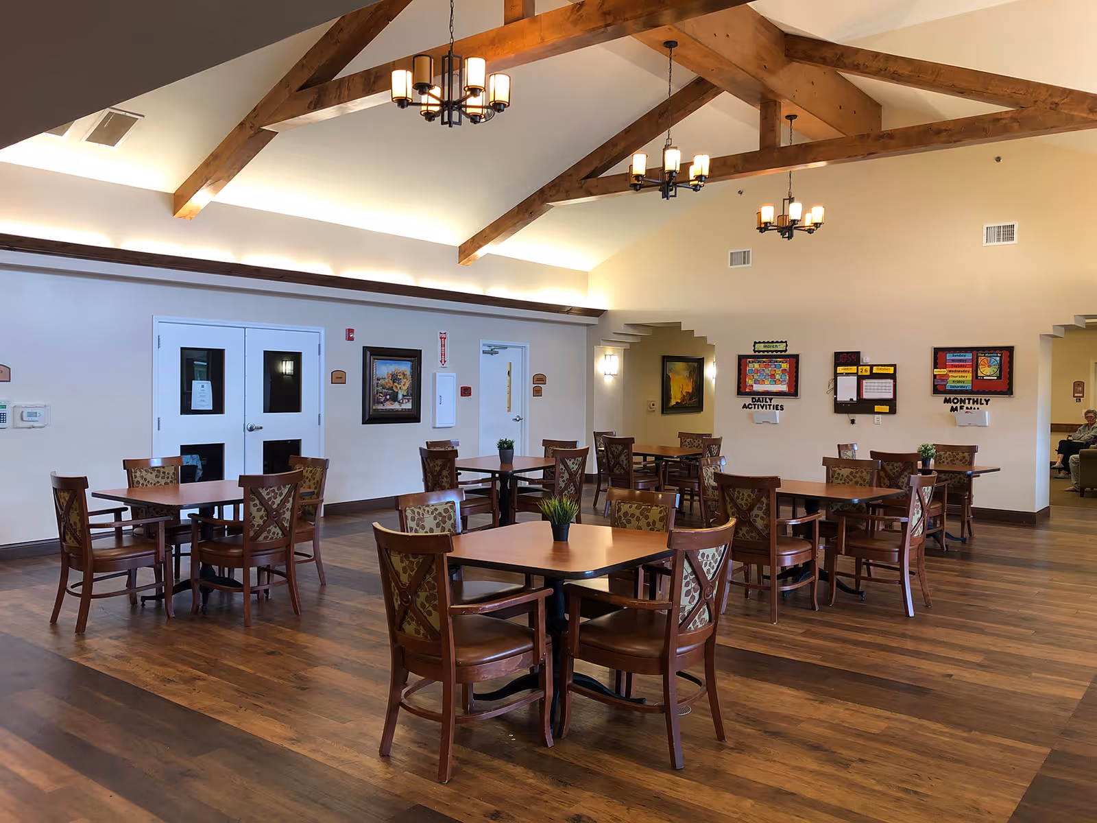 A spacious dining room with wooden floors and high ceilings featuring exposed wooden beams. Several square wooden tables with patterned cushioned chairs are arranged throughout the room. The walls are light-colored with framed artwork and bulletin boards displaying daily activities and monthly menus. There are two white double doors on one side and a few wall-mounted light fixtures. The room is well-lit with hanging chandeliers and natural light.