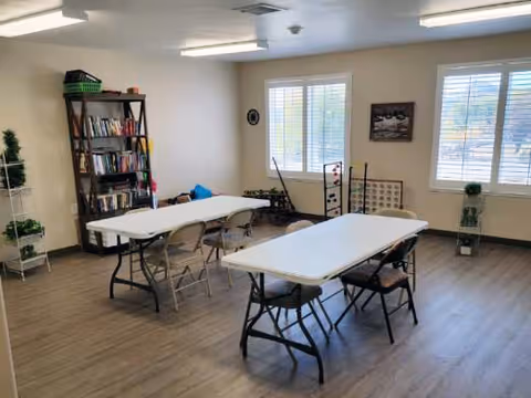 A bright room with two white folding tables and several folding chairs arranged around them. There is a tall bookshelf filled with books and some baskets on the left side. The room has large windows with white blinds letting in natural light, and a few small plants and decorative items are placed around the room.