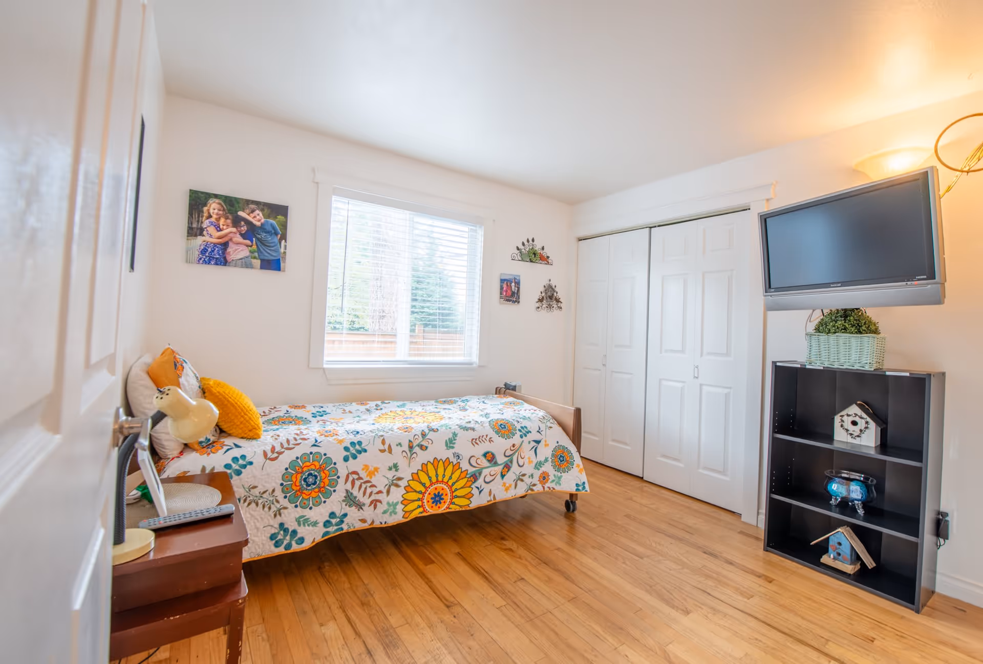 A bright bedroom with a single bed covered in a colorful floral quilt, a wooden floor, a window with white blinds, a wall-mounted TV above a black shelf with decorative items, and a closet with white sliding doors.