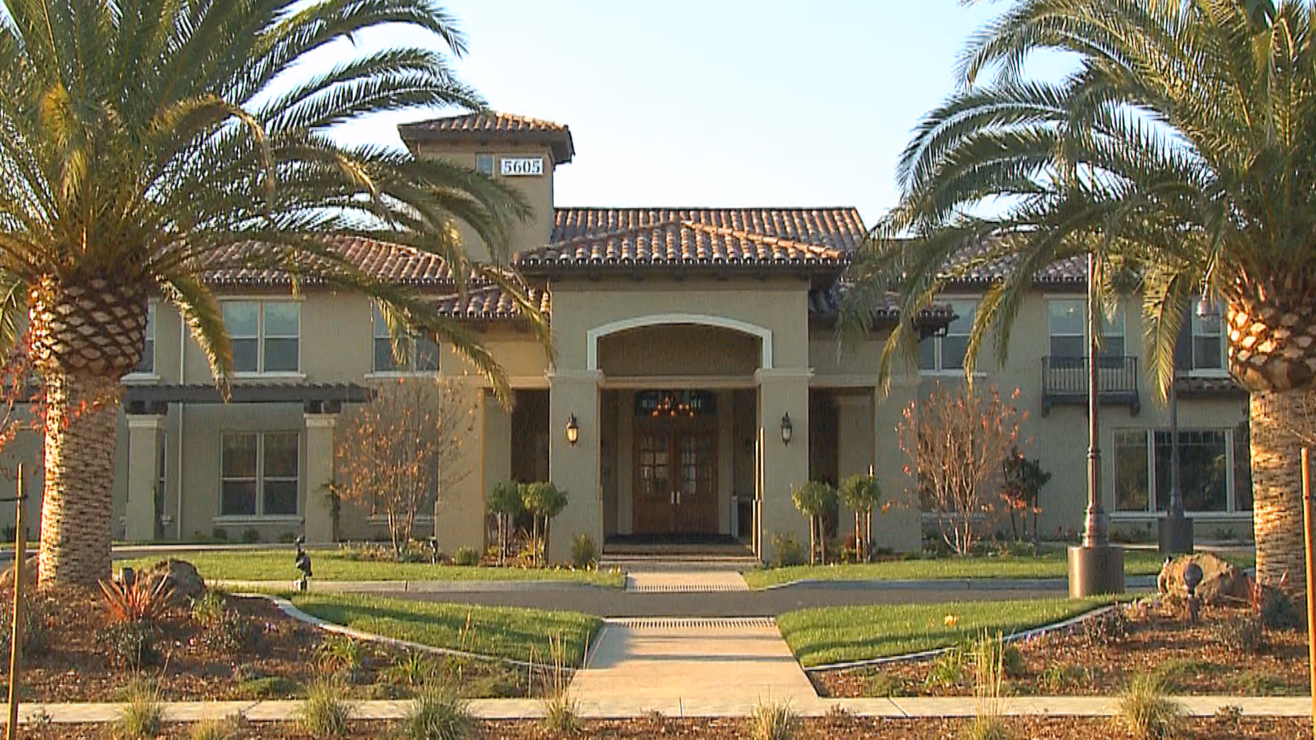 Front exterior view of a two-story building with a tiled roof, beige walls, and a central entrance with an archway. The building is flanked by tall palm trees and landscaped greenery with a concrete walkway leading to the entrance.