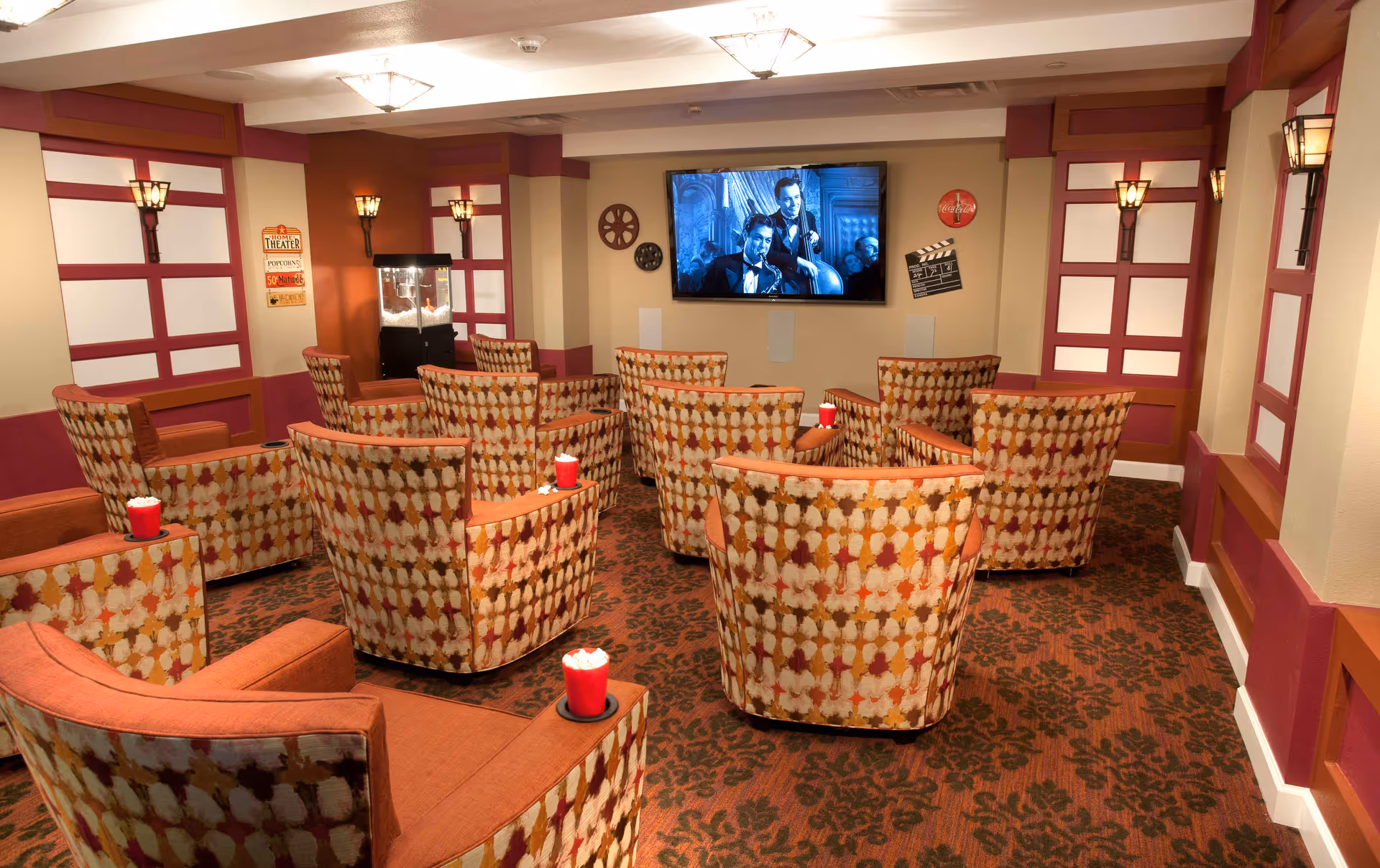 A cozy home theater room with patterned armchairs arranged in rows facing a large flat-screen TV mounted on the wall. The TV is playing a black-and-white movie featuring two men in tuxedos. The room has warm lighting with wall sconces and ceiling lights, a popcorn machine in the corner, and movie-themed decorations on the walls including a film reel, a clapperboard, and a Coca-Cola sign. Each chair has a red cup filled with popcorn in a built-in cup holder.