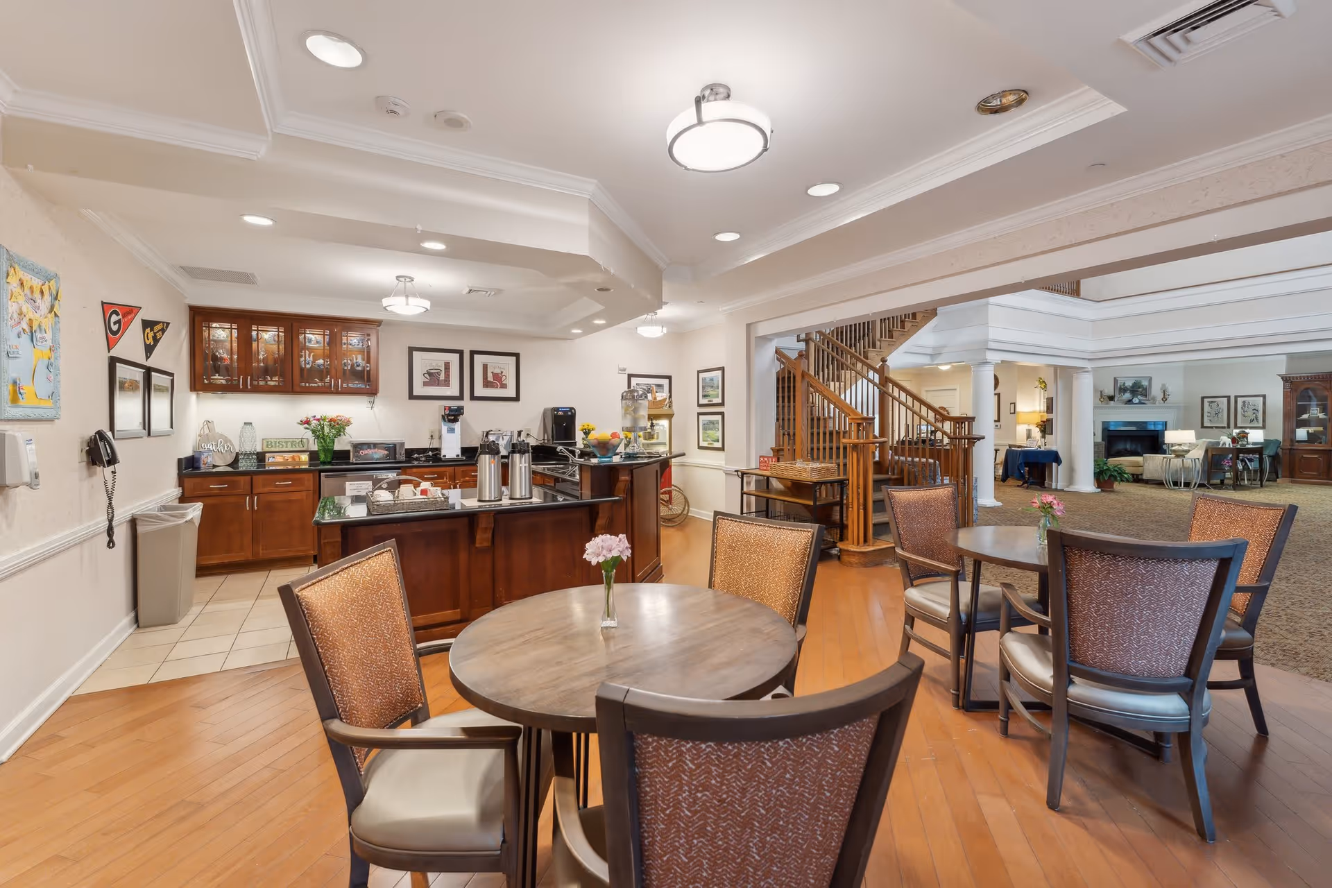 Interior view of a senior living facility common area with a small kitchen or coffee station on the left, featuring wooden cabinets, coffee dispensers, and a countertop with snacks. In the foreground, there are round wooden tables with chairs arranged around them, each table having a small vase with flowers. In the background, there is a wooden staircase leading to an upper floor and a lounge area with carpet, a fireplace, and additional seating.