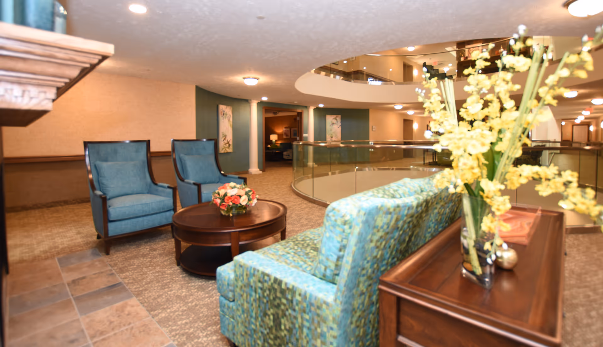 A cozy seating area in a retirement facility featuring two blue upholstered armchairs and a patterned blue sofa arranged around a round wooden coffee table with a floral centerpiece. The space has beige walls, carpeted floors, and a wooden side table with a vase of yellow flowers. The background shows a curved glass railing overlooking a lower level and hallway with warm lighting.
