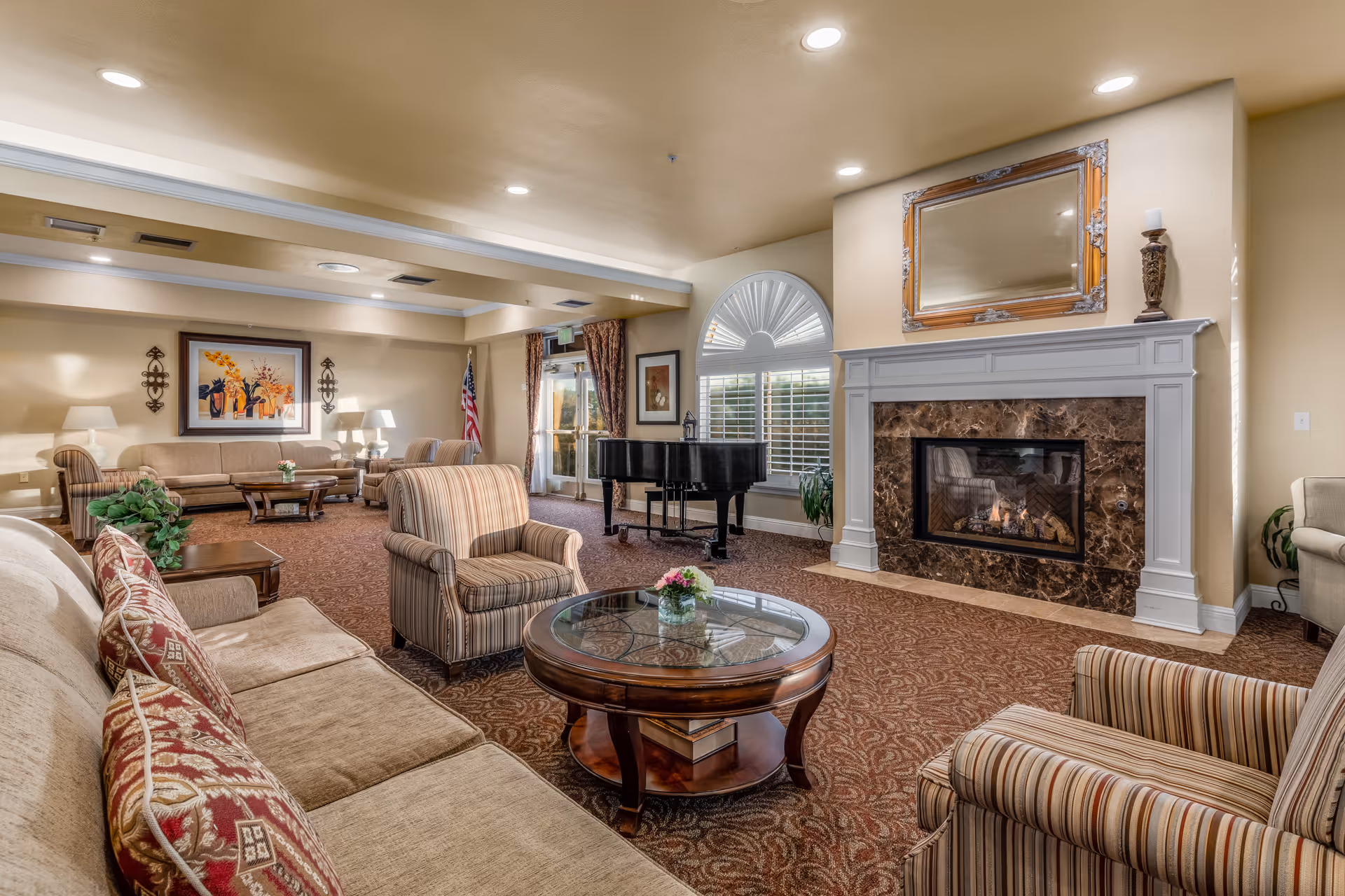 A cozy living room area with beige walls and carpeted floor. The room features a fireplace with a marble surround and a large mirror above it. There are multiple upholstered chairs and sofas arranged around a round glass-top coffee table with a small flower arrangement. A black grand piano is positioned near a window with white plantation shutters and an arched top. The room is softly lit with recessed ceiling lights and table lamps, and there is an American flag near the back wall.