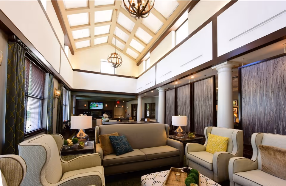 A spacious and well-lit living room area with high vaulted ceilings featuring skylights and hanging light fixtures. The room has comfortable seating including a sofa and armchairs with decorative pillows, side tables with lamps, and large windows with curtains. In the background, there is a television mounted on the wall and a person sitting on a chair.