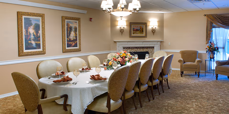 A formal dining room with a long table covered with a white tablecloth, set with plates, glasses, and folded napkins. The table is surrounded by upholstered chairs. The room has beige walls with framed landscape paintings, a fireplace with a small painting above it, and a chandelier hanging from the ceiling. There are also armchairs and a side table with a flower arrangement near a window with sheer curtains.