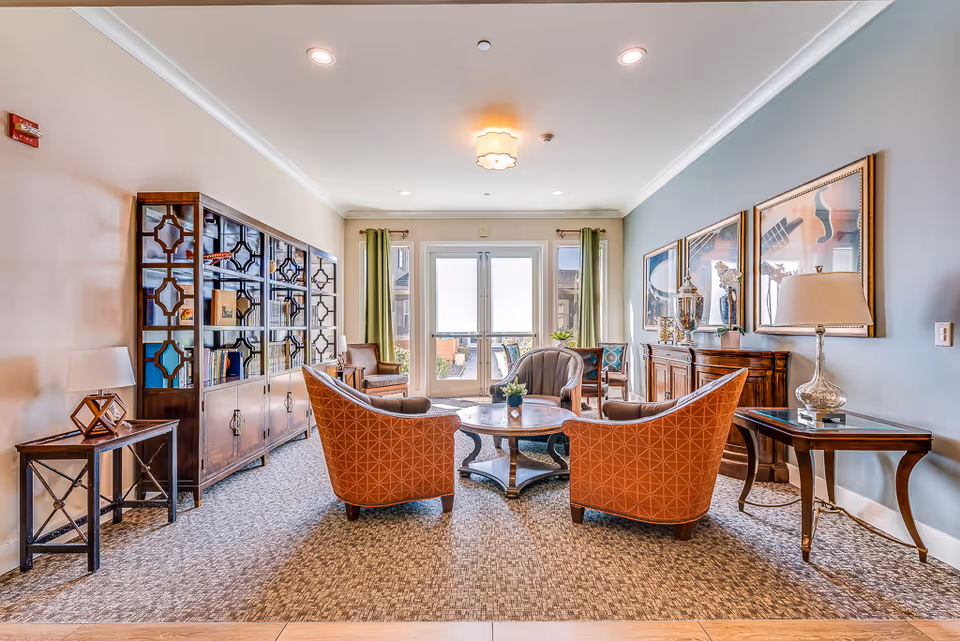 A bright and elegant living room area in a senior living facility featuring four upholstered armchairs arranged around a round wooden coffee table. The room has a large wooden cabinet with glass doors on the left, a side table with a lamp, and a wooden console table with decorative items and framed artwork on the right wall. Double glass doors with green curtains lead to an outdoor view.