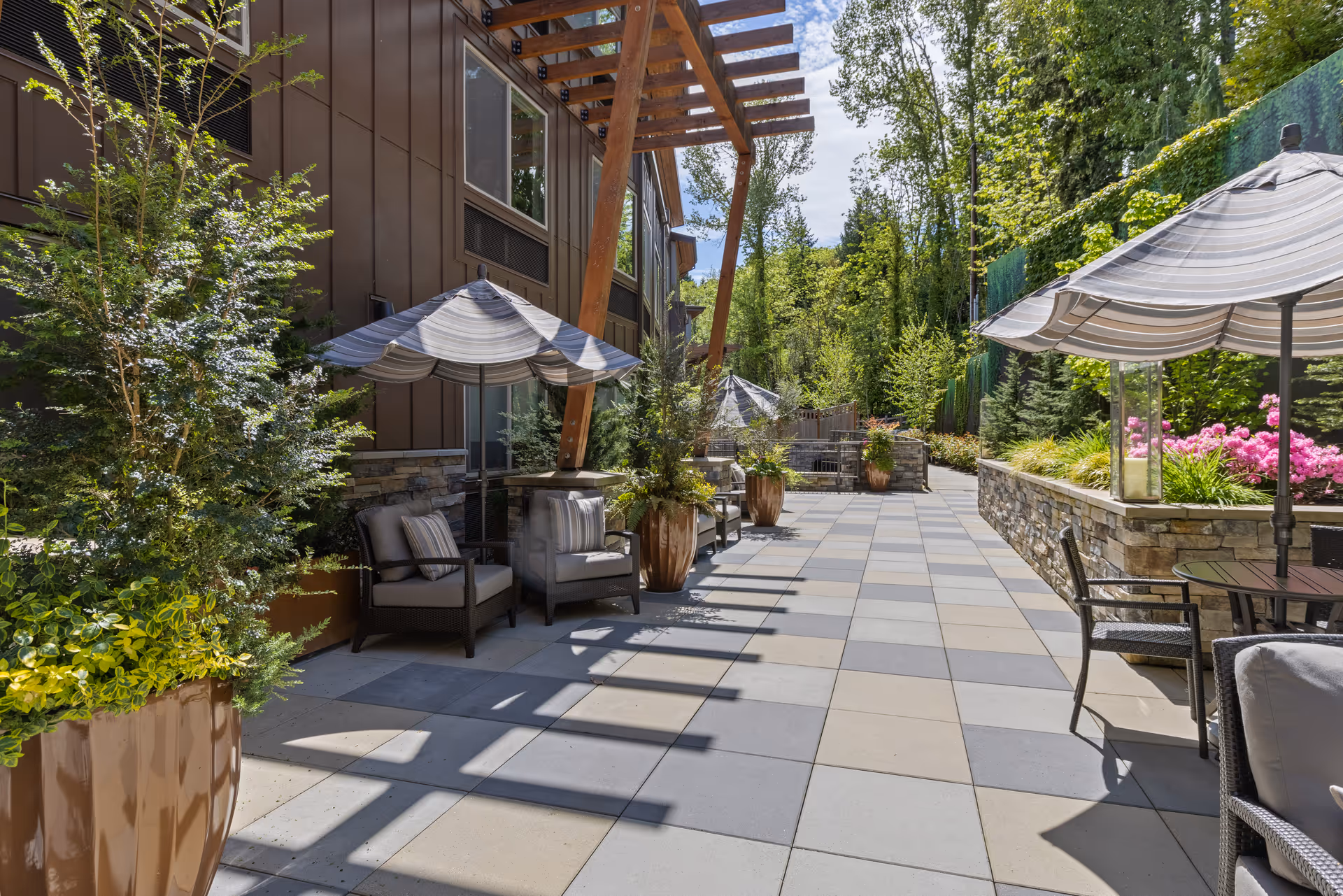 Outdoor patio area at Aegis Living Mercer Island featuring cushioned chairs with striped umbrellas, large potted plants, a stone wall planter with pink flowers, and a tiled floor. The space is surrounded by trees and greenery under a partly cloudy sky.