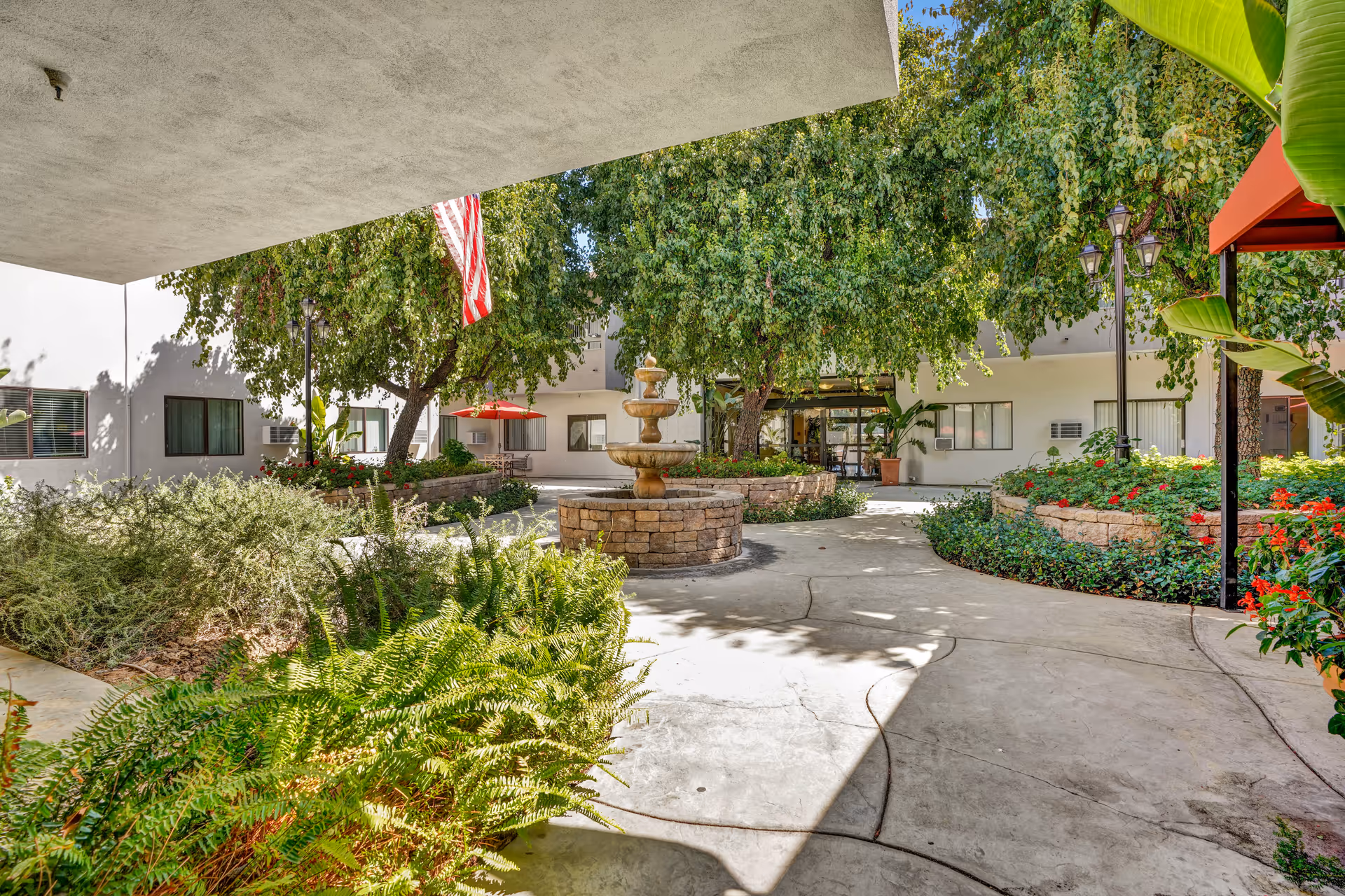 Outdoor courtyard area at Valley Silvertown featuring a central stone fountain, surrounded by lush greenery, trees, and flower beds. There are paved walkways, an American flag hanging from a tree, and a building with windows in the background.