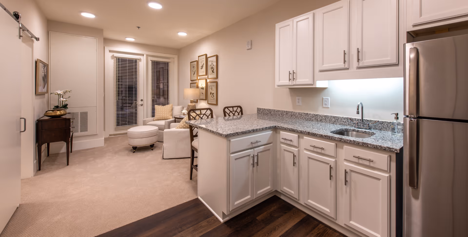 Interior view of a senior living facility unit featuring a modern kitchen with white cabinets, granite countertops, a stainless steel refrigerator, and a sink. Adjacent to the kitchen is a cozy living area with a white armchair, ottoman, side table with a lamp, and framed artwork on the wall. The room has beige carpeting and wooden flooring near the kitchen, with a door and windows with blinds in the background.