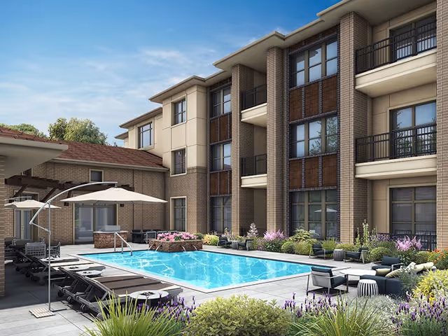 Outdoor courtyard area of a senior living facility with a swimming pool surrounded by lounge chairs, umbrellas, and various plants and flowers. The building has multiple floors with balconies overlooking the pool area.