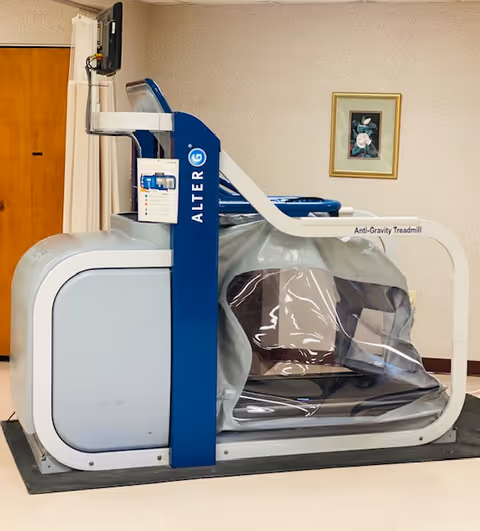 An AlterG Anti-Gravity Treadmill in a room with beige walls and a wooden door. The treadmill has a transparent plastic enclosure and a blue and white frame. A framed floral picture hangs on the wall behind the treadmill.