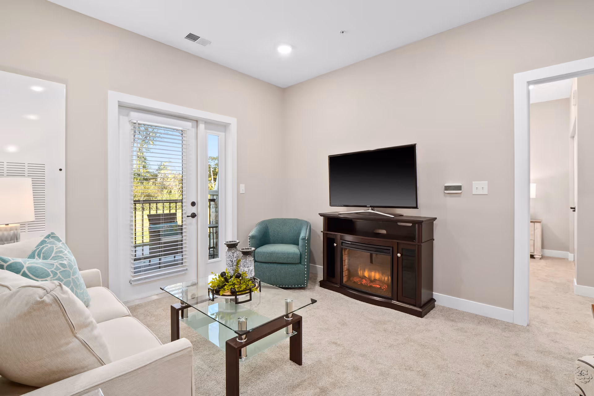 A cozy living room with a beige sofa adorned with blue patterned pillows, a glass coffee table with decorative items, a teal armchair, and a dark wooden TV stand with an electric fireplace. There is a flat-screen TV on the stand and a door with blinds leading to an outdoor area with greenery visible outside.