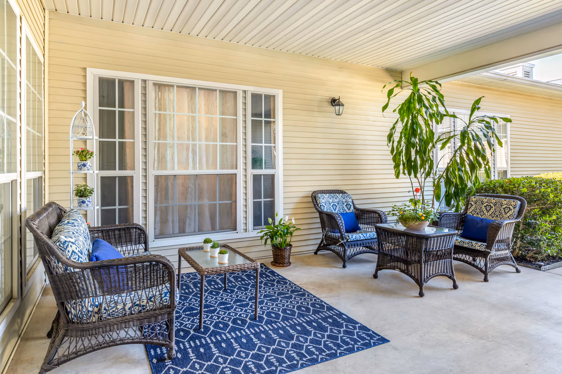 Covered outdoor patio area with wicker furniture including a loveseat, two armchairs, and two glass-top tables. The furniture has blue and white patterned cushions and blue throw pillows. There is a blue patterned rug on the concrete floor, several potted plants, and beige siding on the building walls with large windows and a wall-mounted light fixture.