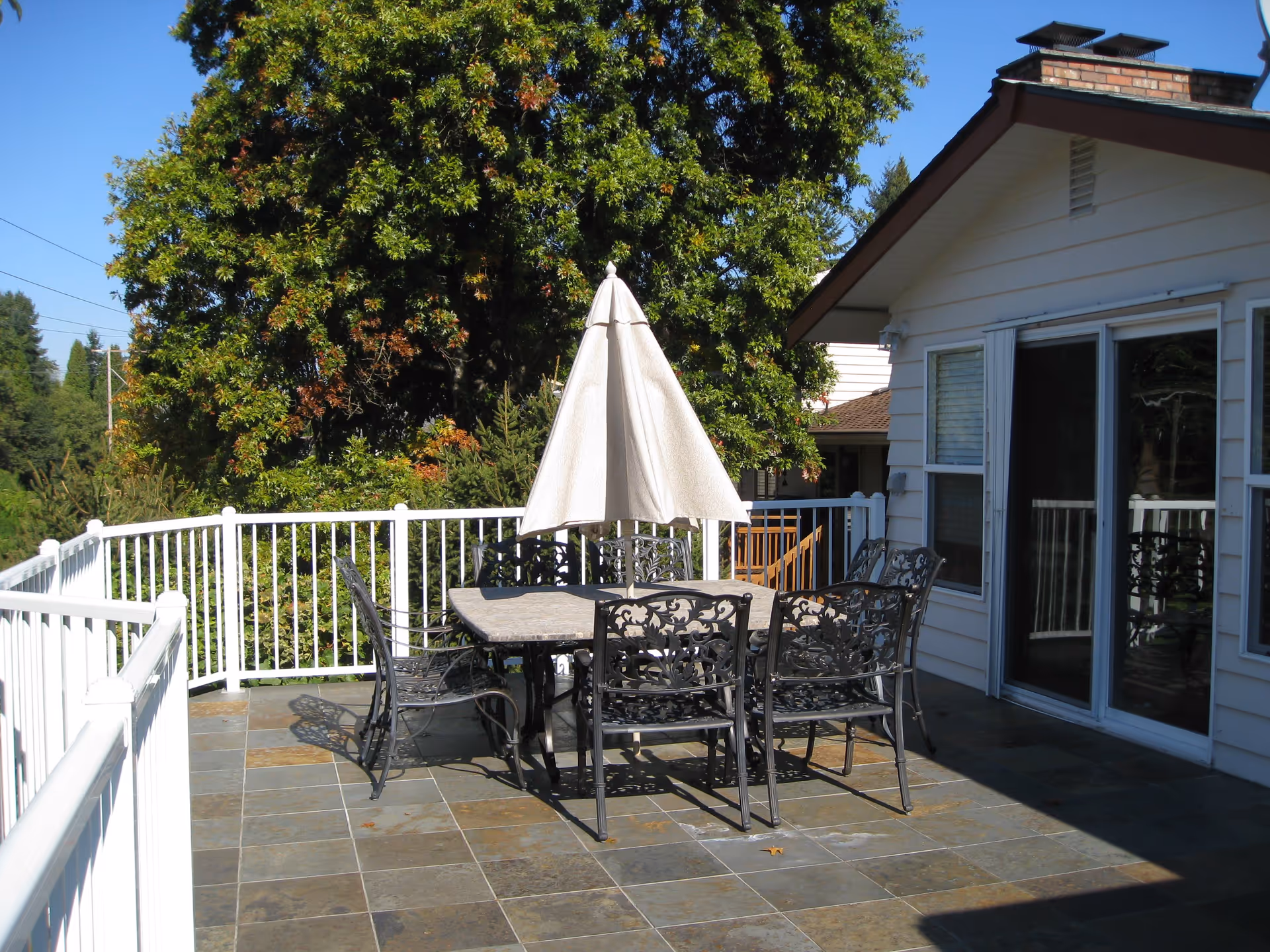 Outdoor patio area with a tiled floor, a metal table with six matching chairs, and a closed beige umbrella in the center. The patio is surrounded by a white railing and adjacent to a house with sliding glass doors and windows. Large green trees are visible in the background under a clear blue sky.