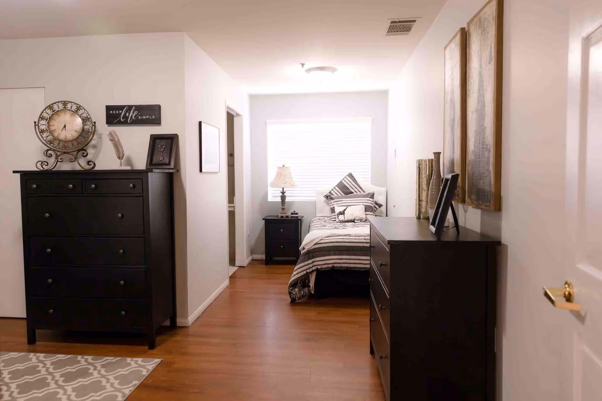 A cozy bedroom with wooden flooring, featuring a single bed with striped bedding and multiple pillows near a window with blinds. There are two black dressers, one on the left with a decorative clock and framed art, and another on the right with vases and framed pictures. The walls are light-colored, and there is a small nightstand with a lamp next to the bed.