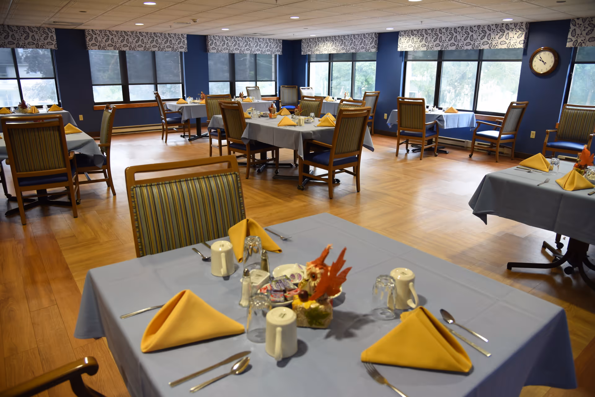 A dining room in a senior living facility with multiple tables covered in light blue tablecloths and set with yellow folded napkins, silverware, cups, and small centerpieces. The room has large windows with patterned valances, blue walls, wooden flooring, and several wooden chairs with striped cushions. A clock is mounted on the wall.