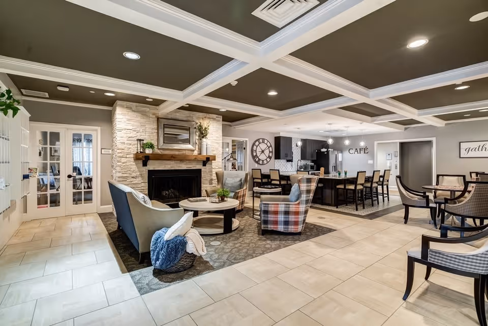 A cozy and modern common area in a senior living facility featuring a stone fireplace with a wooden mantel, comfortable seating including a sofa and plaid armchairs, a round coffee table, and a basket with blankets. In the background, there is a café area with a large island, bar stools, pendant lighting, and a wall clock. The room has a coffered ceiling with recessed lighting and light-colored tiled flooring.
