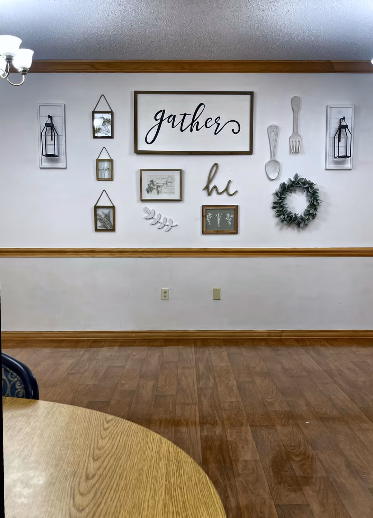 Interior wall decorated with framed pictures, a wreath, and wooden utensils. The largest frame has the word 'gather' written in cursive. The wall has a wooden chair rail and light-colored paint. Part of a round wooden table and a chandelier are visible.