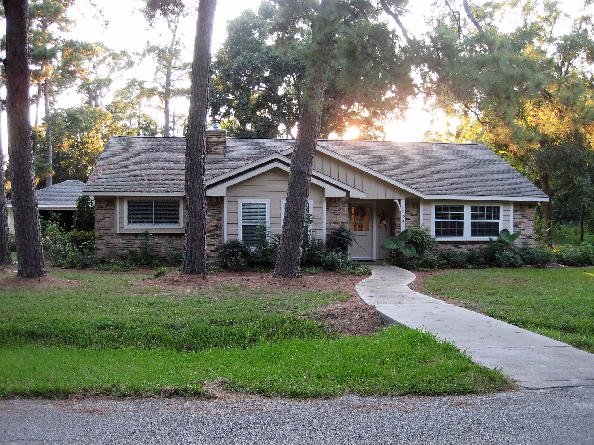 Single-story house with a brick and beige exterior surrounded by trees and greenery. A curved concrete walkway leads from the street to the front door. The house has multiple windows and a shingled roof, with sunlight filtering through the trees in the background.