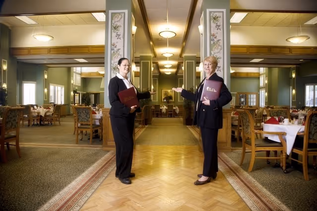 Two women dressed in black suits standing in a spacious dining room with tables and chairs arranged neatly. They are each holding a menu and gesturing towards the dining area, which features green walls, large mirrors, and soft lighting fixtures.