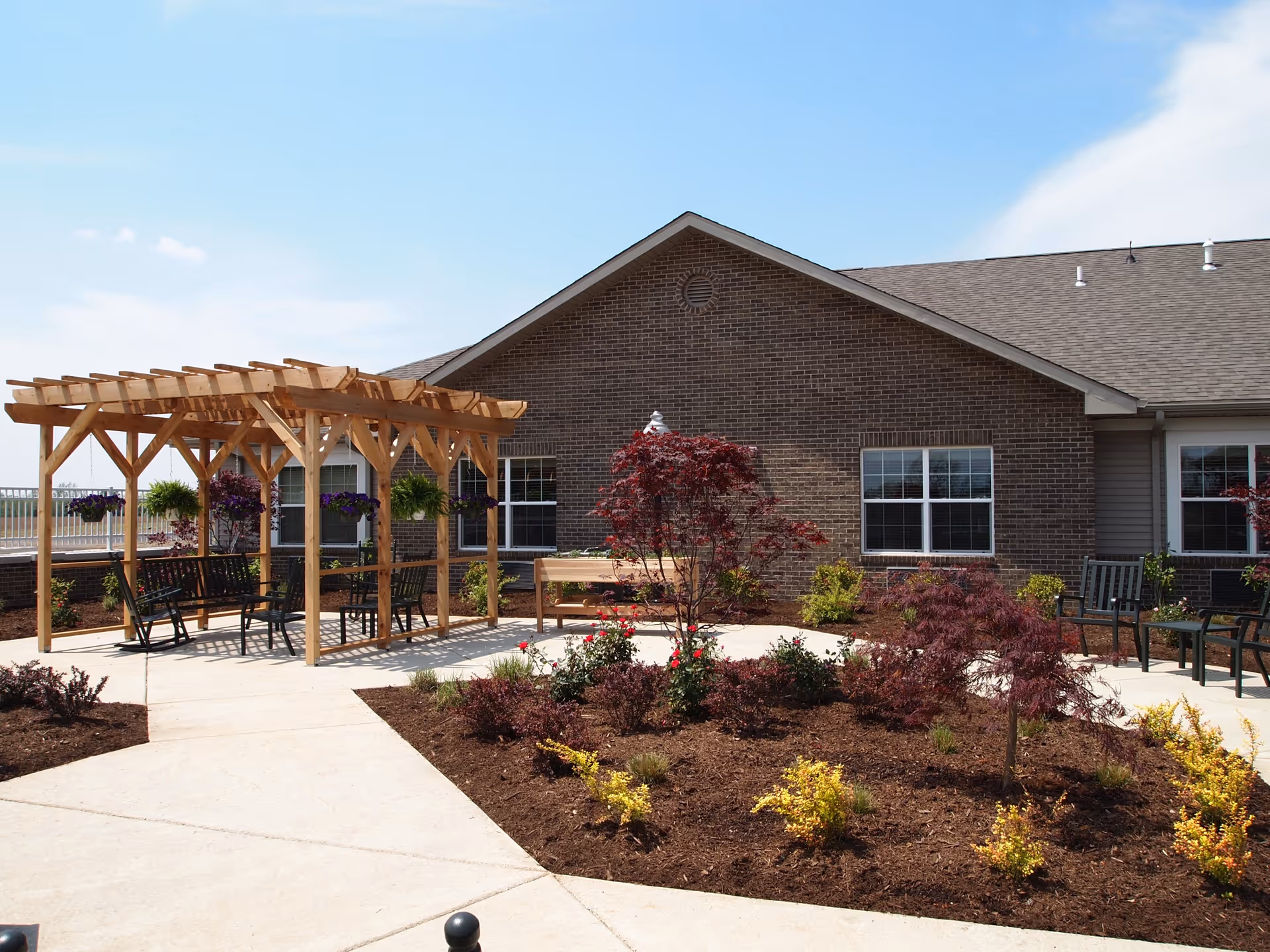 Outdoor patio area at Blair Ridge Health Campus featuring a wooden pergola with hanging plants and benches underneath. The patio is surrounded by landscaped garden beds with small trees and shrubs, and the building's brick exterior is visible in the background under a partly cloudy sky.