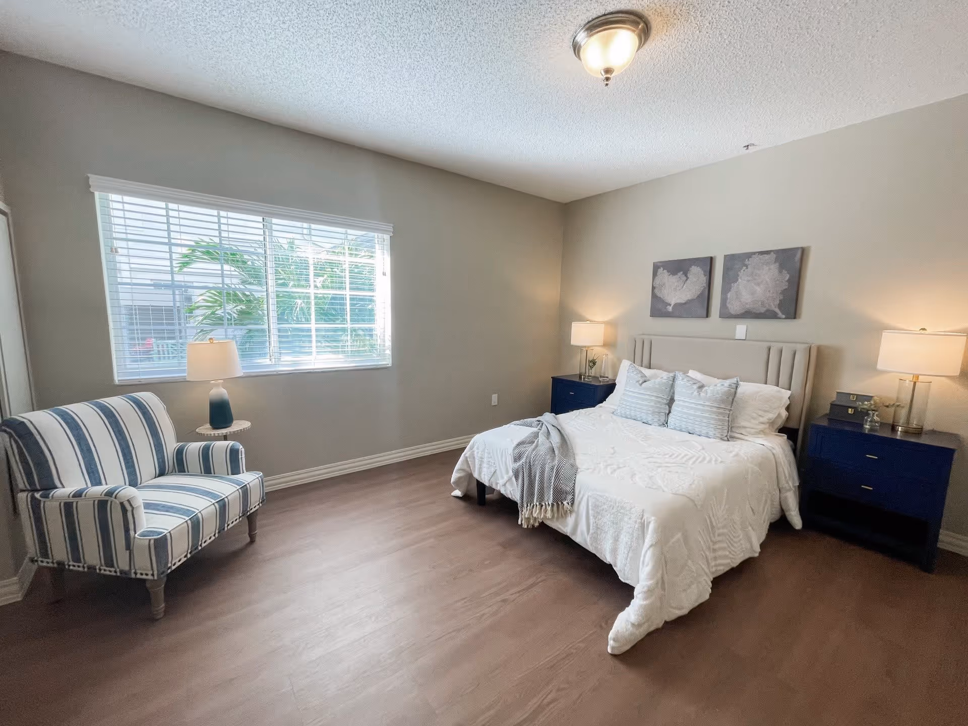 Well-lit bedroom with a neatly made bed, two navy nightstands with lamps, and a striped armchair by a window.
