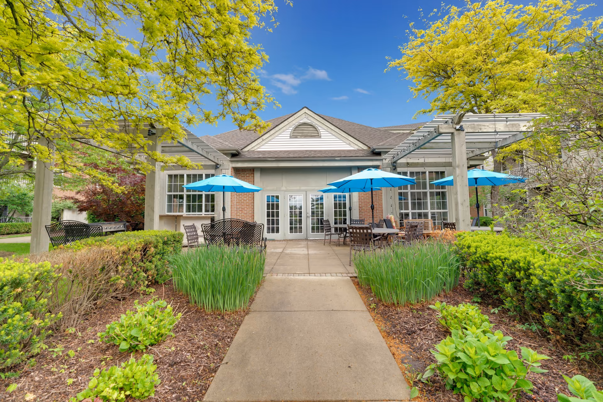Outdoor patio area at Shorehaven facility with a concrete walkway leading to a building entrance. The patio features tables with blue umbrellas, metal chairs, and benches surrounded by green shrubs and trees under a clear blue sky.