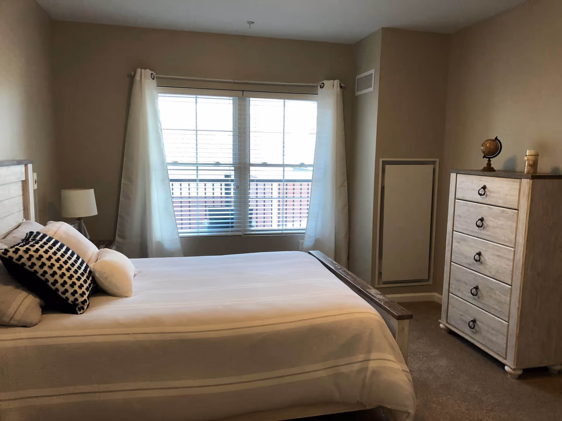 A bedroom with a neatly made bed featuring white and patterned pillows, a wooden headboard, a bedside lamp, a window with white curtains and blinds, and a wooden dresser with a small globe and decorative items on top.