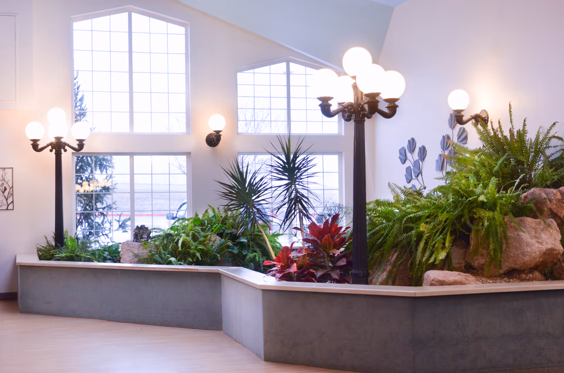 Bright indoor atrium with large arched windows, lush plants, rocks and decorative lamp posts.