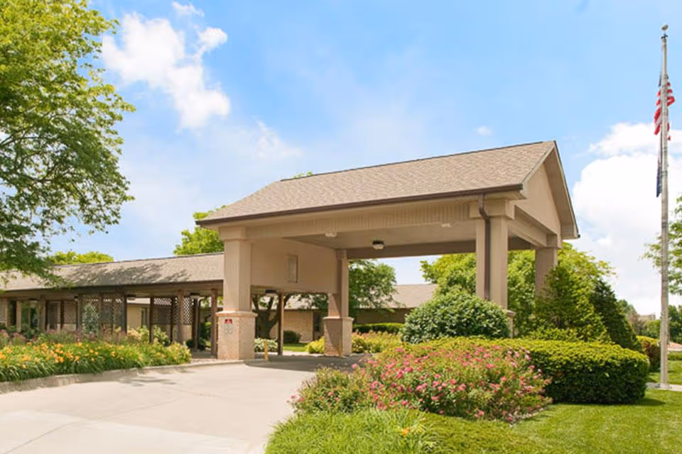 Entrance area of a senior living facility with a covered drop-off zone, surrounded by well-maintained greenery, bushes, and flowering plants under a blue sky with some clouds.